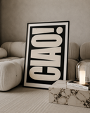 A large framed black and white art print with the word 'CIAO!' in bold letters, leaning against a beige sofa in a modern living room setting.