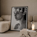 A framed black-and-white Vogue magazine cover print leaning against a beige sofa in a styled living room setting, with a marble side table, lit glass candle holder, and book nearby.